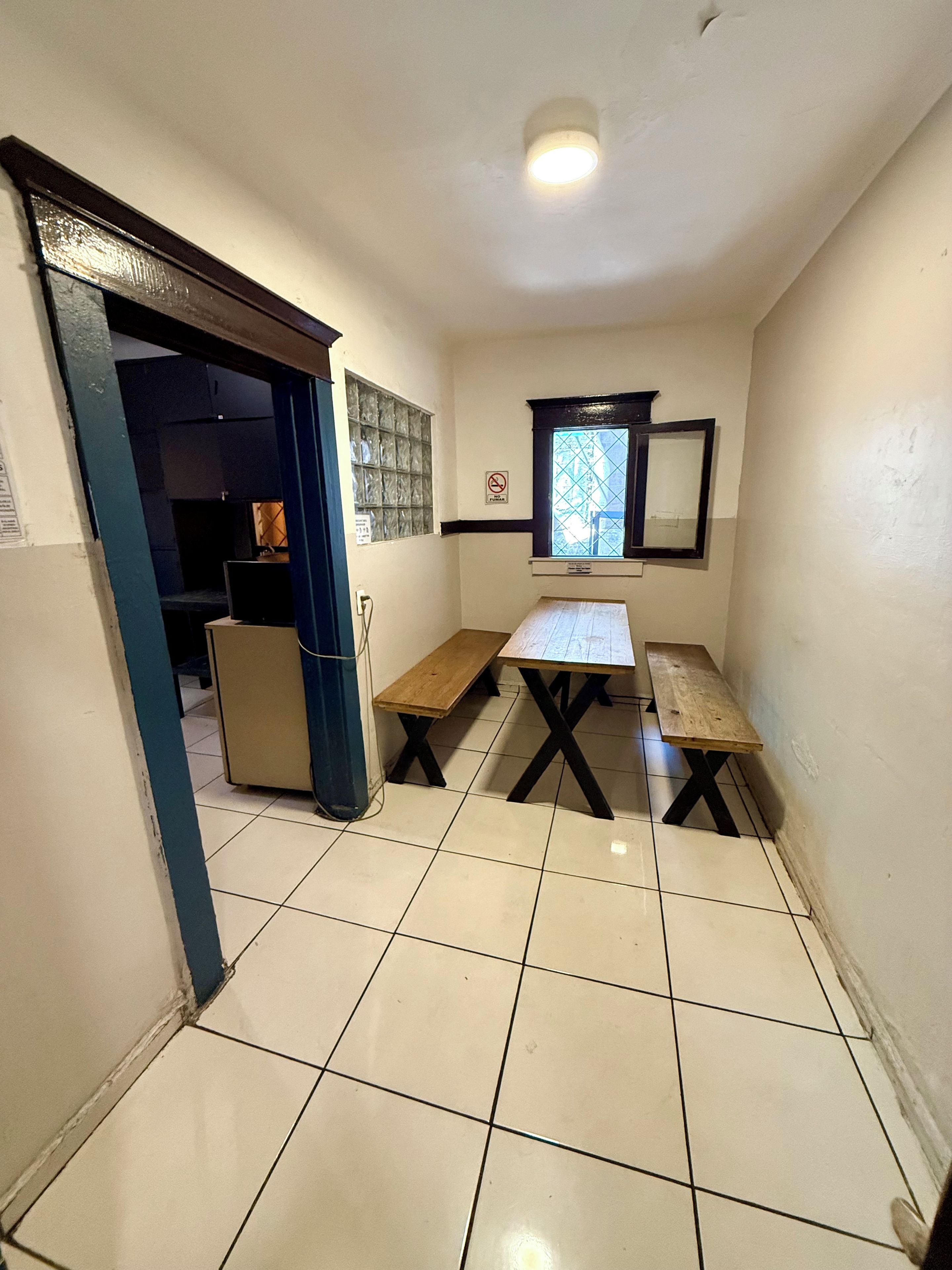 A small, narrow room with a tiled floor, a wooden bench, a table, a window with a view, and a door leading to another room.