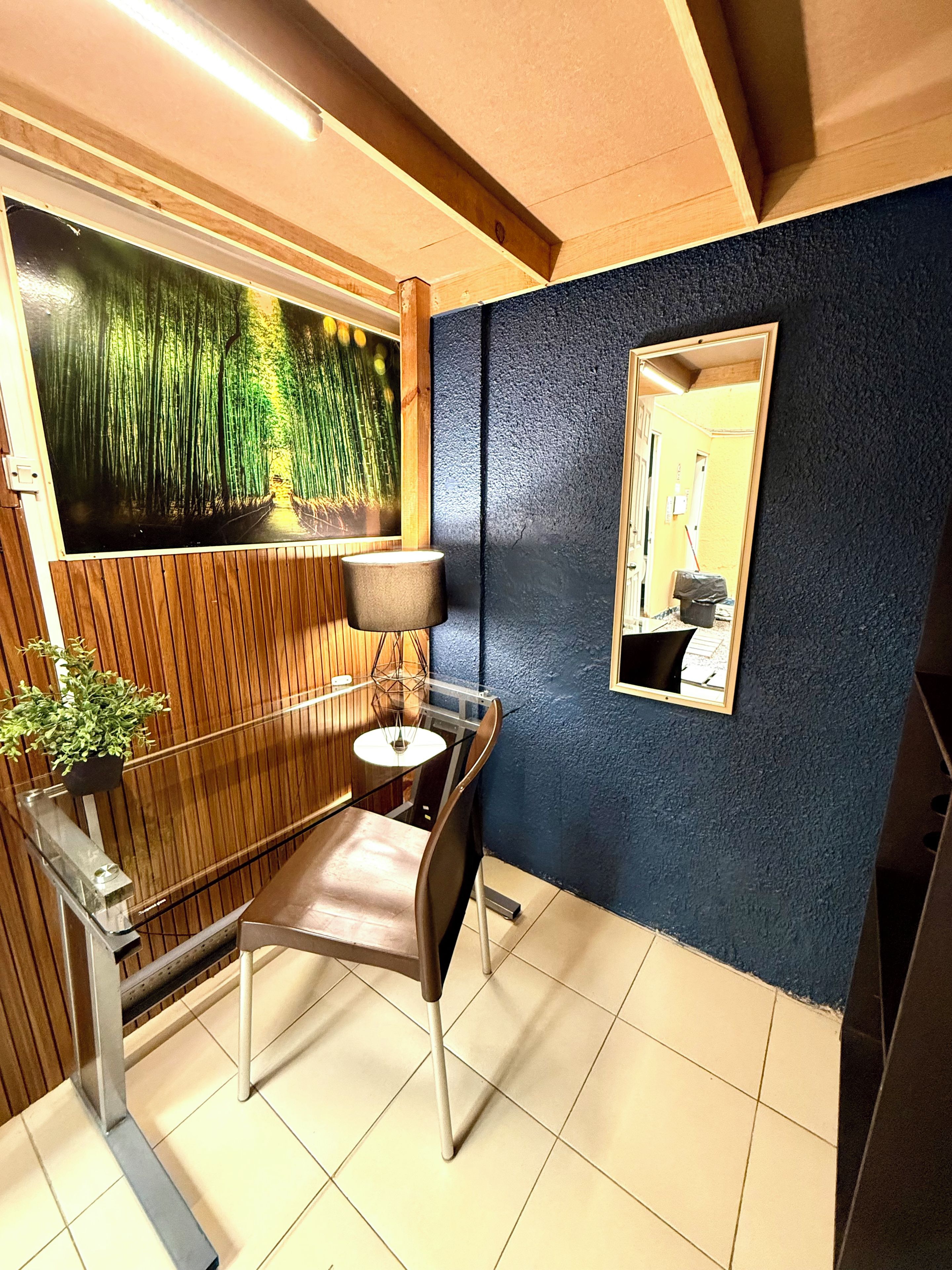 Een kamer met een houten vloer, een stoel, een tafel, een lamp, een wandspiegel en een groot schilderij van een bos.