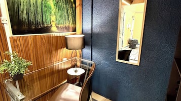 Una habitación con piso de madera, una silla, una mesa, una lámpara, un espejo en la pared y un cuadro grande de un bosque.