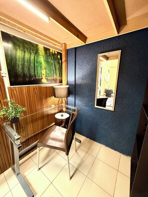 Ett rum med trägolv, en stol, ett bord, en lampa, en spegel som hänger på väggen och en stor målning som föreställer en skog.