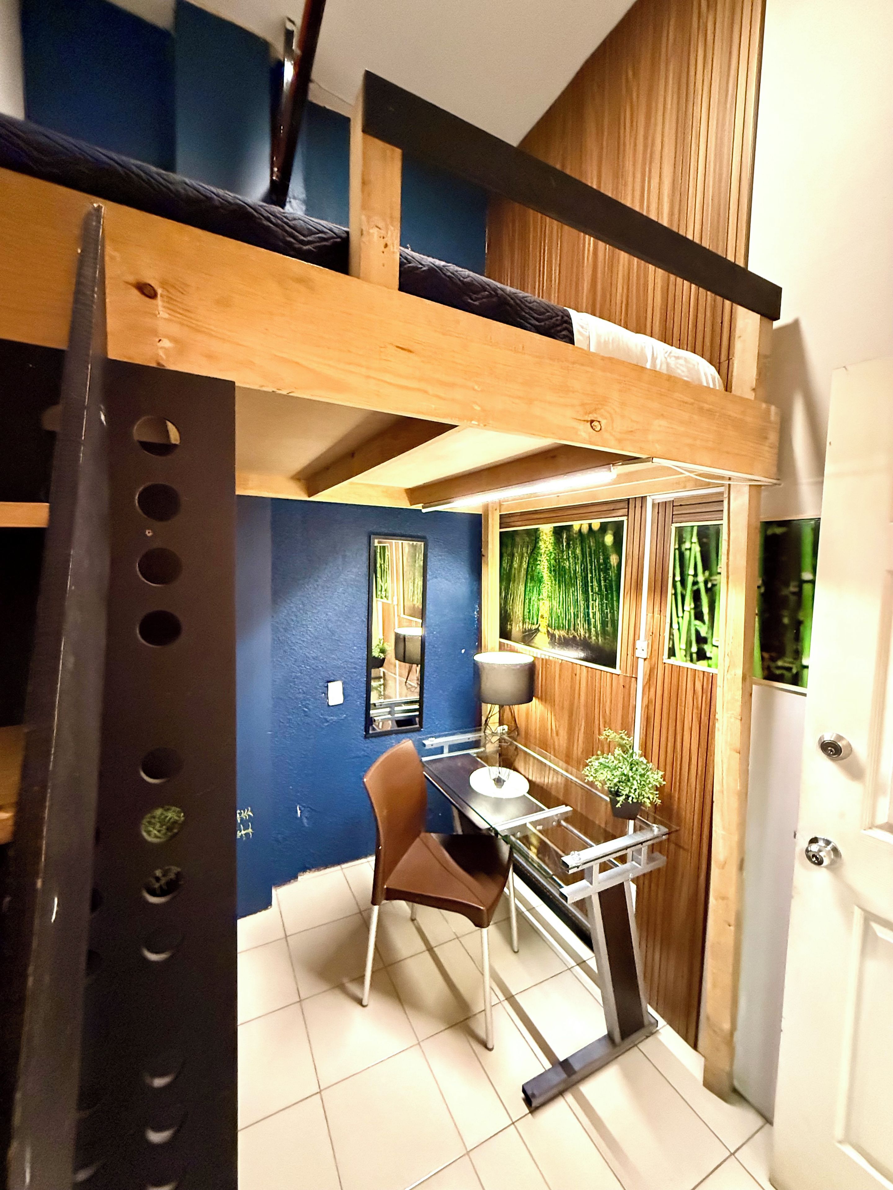 Habitación pequeña con literas, una mesita con una silla, una planta en maceta y ventana con vistas a bambú.