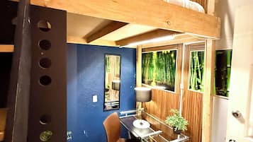 Habitación pequeña con literas, una mesita con una silla, una planta en maceta y ventana con vistas a bambú.