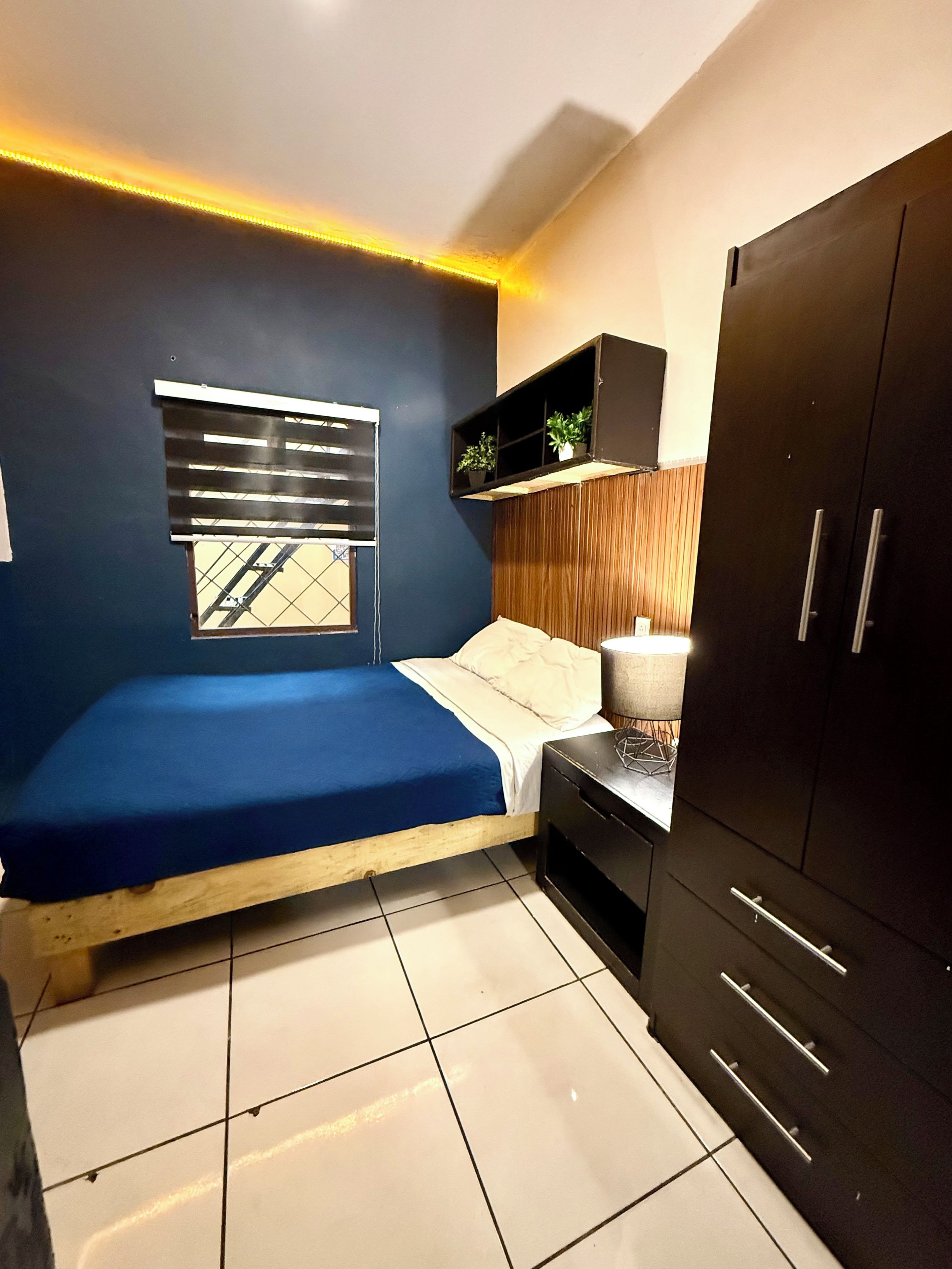 Un dormitorio con cama, ventana con persianas, mesita de noche con lámpara, armario y estante con plantas.