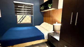 Un dormitorio con cama, ventana con persianas, mesita de noche con lámpara, armario y estante con plantas.