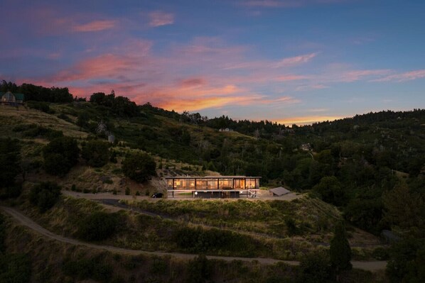 A modern house with a glass facade, surrounded by a hilly landscape.