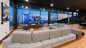 En moderne stue med en stor grå hjørnesofa, et fladskærms-tv og store vinduer med udsigt over byen.