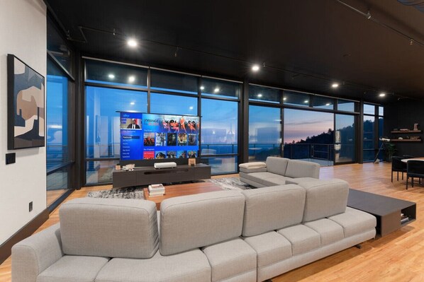 Una sala de estar moderna con un amplio sofá seccional gris, un televisor de pantalla plana y ventanas grandes con vistas a la ciudad.