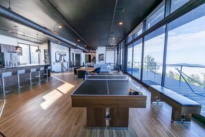 Una habitación amplia con mesa de ping pong, taburetes de bar y un gran ventanal con vista al mar.