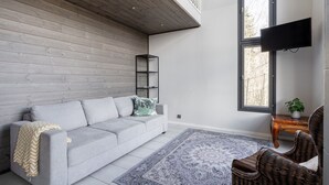 A modern living room with a grey sofa, a patterned rug, a wicker chair, a wooden ceiling, and a large window. - Spacious & Cozy 2BR Cabin at Riihivuori Peak next to Ski Slopes (Muurame)