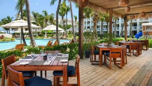 A wooden deck with tables and chairs, a pool, palm trees, and a building in the background. - Deluxe 1 BR, All-Inclusive Suite at Alexandra Resort (The Bight Settlement)