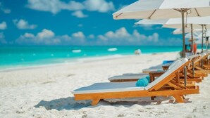 Une plage de sable blanc bordée d’une eau turquoise. On y trouve des chaises longues en bois et des parasols blancs.
