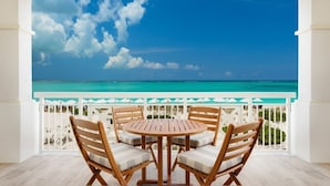 Un balcon avec une table ronde en bois et quatre chaises, d’où l’on peut admirer une mer turquoise et un ciel dégagé.