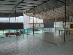Un espacio interior amplio con suelo de baldosas, una zona de estar cubierta y vistas a través de amplias ventanas.
