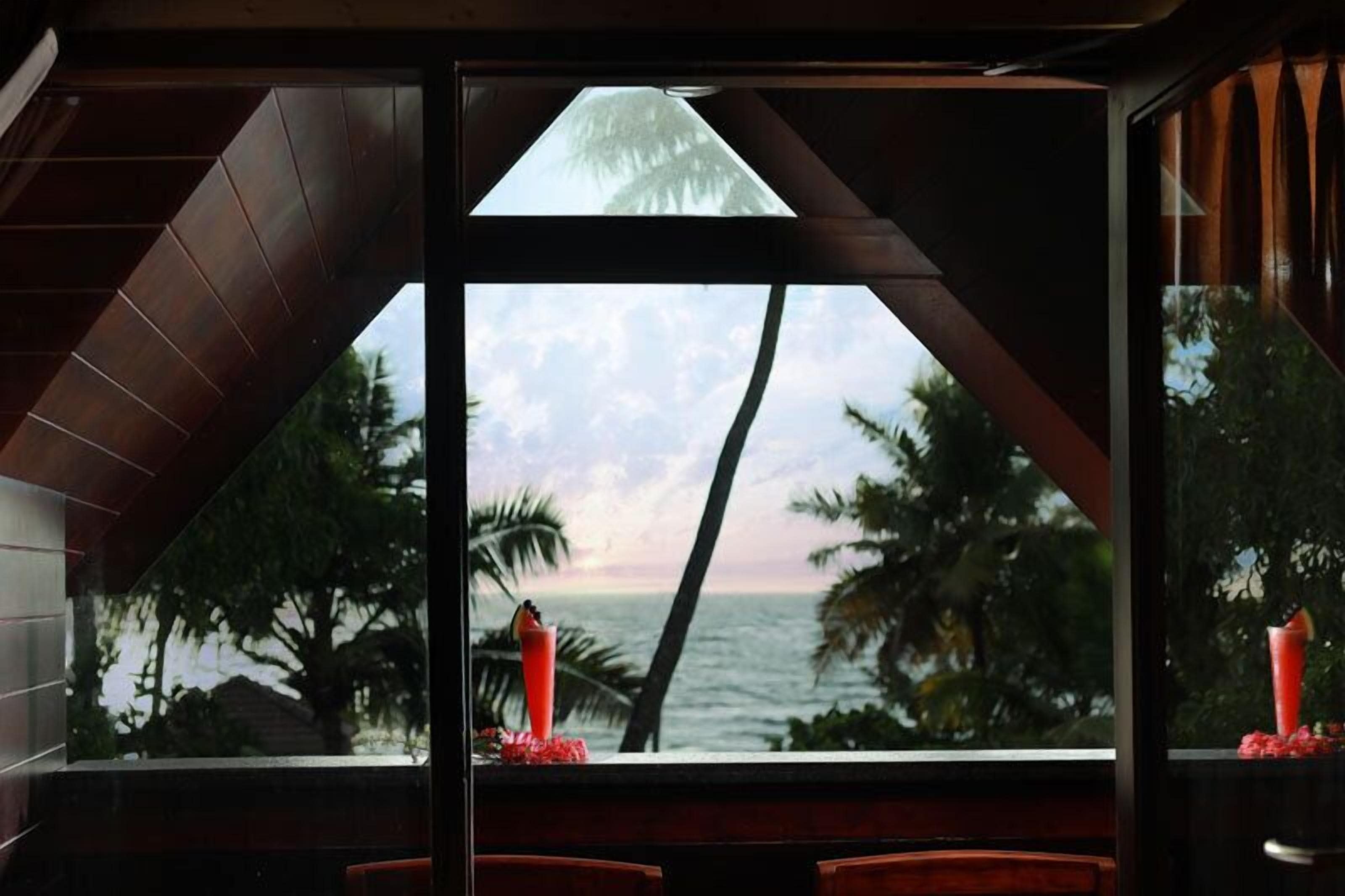 A room with a view of the ocean and palm trees, featuring a large window and a red candle on the windowsill.