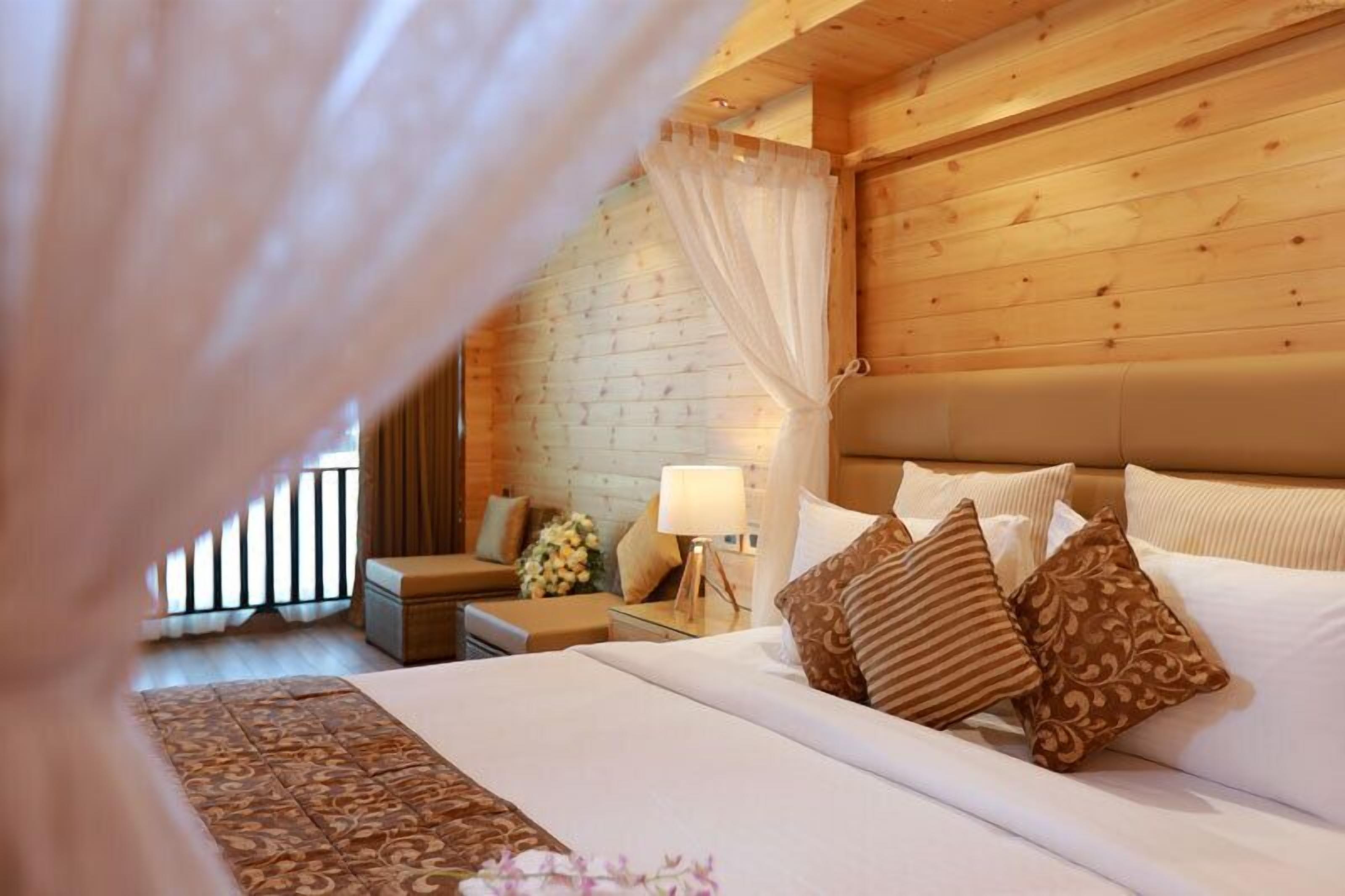 A cozy bedroom with a wooden ceiling, a large bed with patterned pillows, a nightstand with a lamp, and a window with curtains.