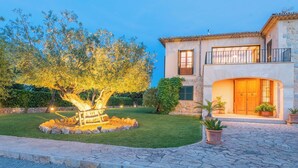 A two-story house with a stone facade, a balcony with a railing, and a well-maintained garden with a tree illuminated by lights.