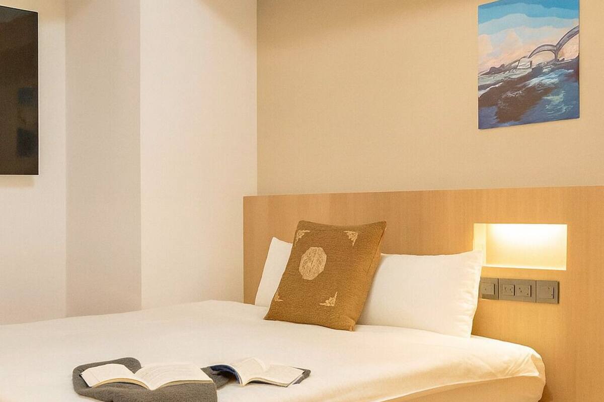Een netjes opgemaakt bed met een bruin kussen, een wit kussen en een grijze plaid. Daarboven hangt een schilderij aan de muur.