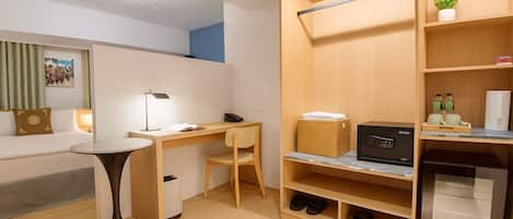 A compact hotel room with a desk, chair, and a small table. There is a wardrobe with shelves and a built-in refrigerator.