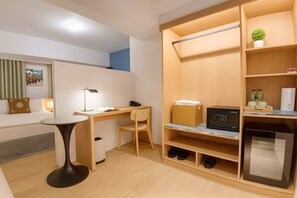 Une chambre d’hôtel compacte comprenant un bureau, une chaise et une petite table. Elle est équipée d’une armoire avec des étagères et d’un réfrigérateur intégré.