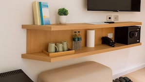 Un salon moderne avec un téléviseur à écran plat fixé au mur, une étagère en bois avec des livres, une petite plante et une machine à café. On y trouve également un pouf beige et un haut-parleur noir posés sur le sol.