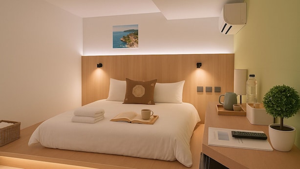 Une chambre d’hôtel moderne avec un lit bien fait, une petite table de chevet avec une lampe, une plante en pot et une vue sur la mer par la fenêtre.