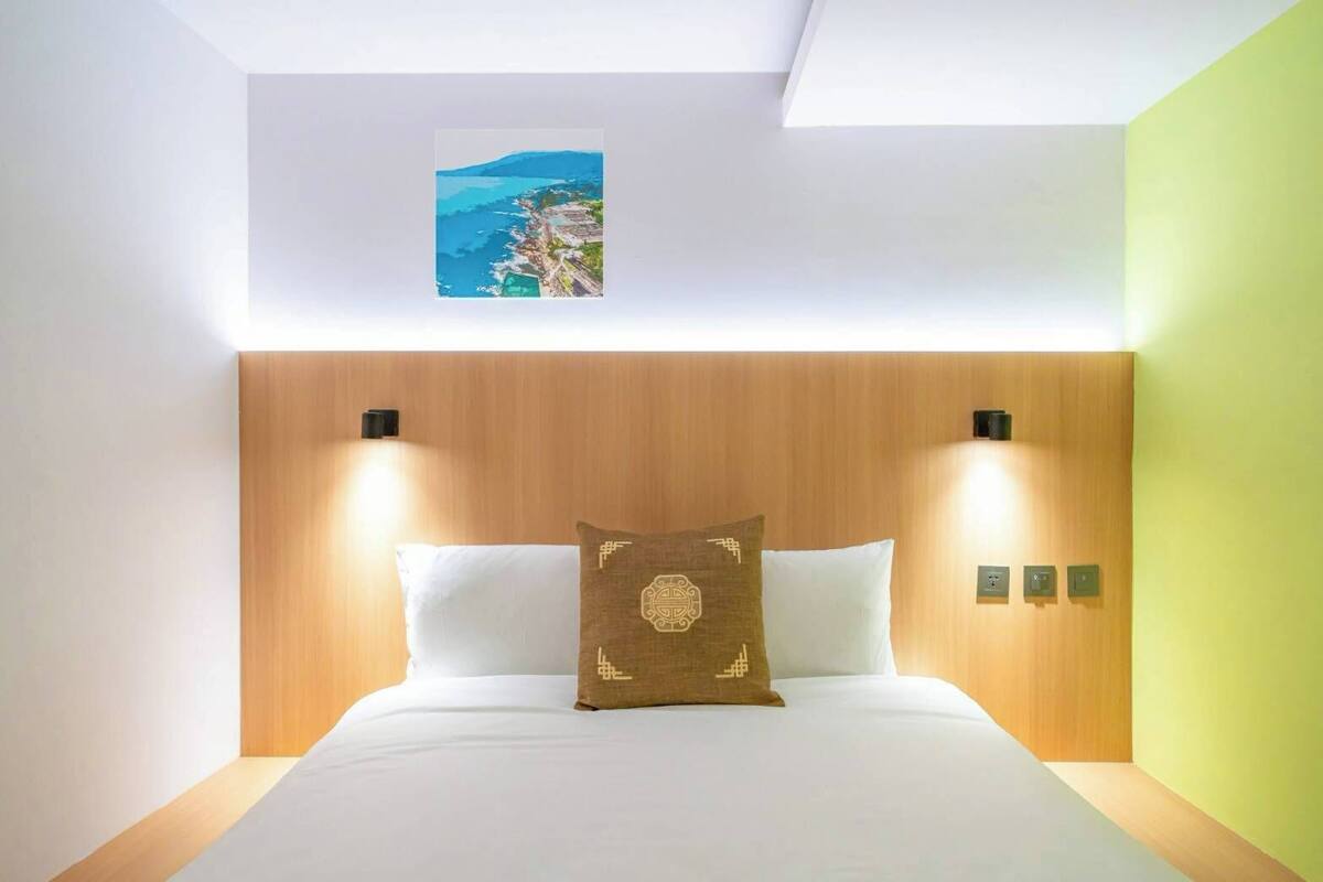 Hotelkamer met houten hoofdeinde, twee bedlampjes, een bed met wit beddengoed en een schilderij van een kustlandschap erboven.