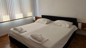 Una cama doble bien tendida con sábanas blancas y dos toallas, una mesita de noche con lámpara, y una ventana con persianas.