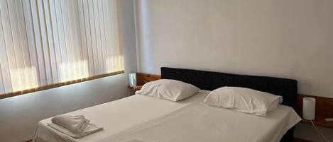 Una cama doble bien tendida con sábanas blancas y dos toallas, una mesita de noche con lámpara, y una ventana con persianas.