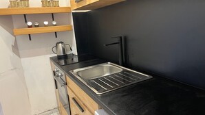 Cocina compacta con encimera negra, pileta de acero inoxidable y hervidor. Hay alacenas de madera y una pequeña repisa con cestas.