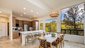 A spacious kitchen with wooden cabinets, a large island, and a dining area with a view of trees and mountains. - Enchanted Waters Retreat (Sedona)