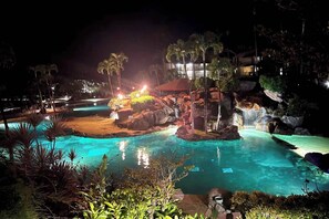 A nighttime resort pool area with clear water, palm trees, and a waterfall feature.