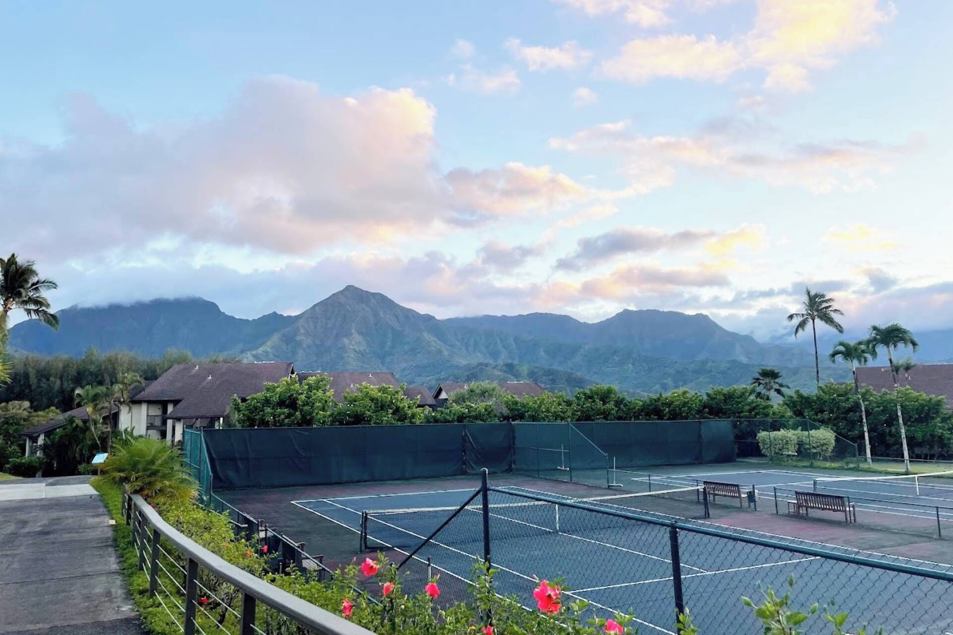 A tennis court with a net, surrounded by a fence and greenery, with mountains in the background.