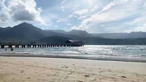Une plage avec une jetée en bois s’avançant dans des eaux calmes, une chaîne de montagnes en arrière-plan et un ciel dégagé.