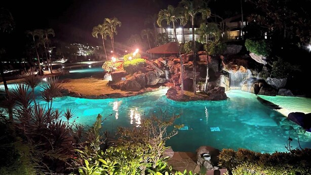 Un espace piscine d’un complexe hôtelier, aménagé pour la nuit, avec une eau cristalline, des palmiers et une cascade.