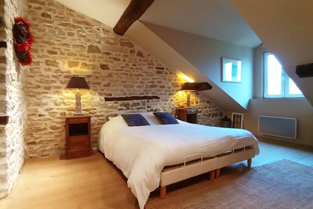 A bedroom with a stone wall, a sloped ceiling, a bed with white bedding, two bedside lamps, and a window.