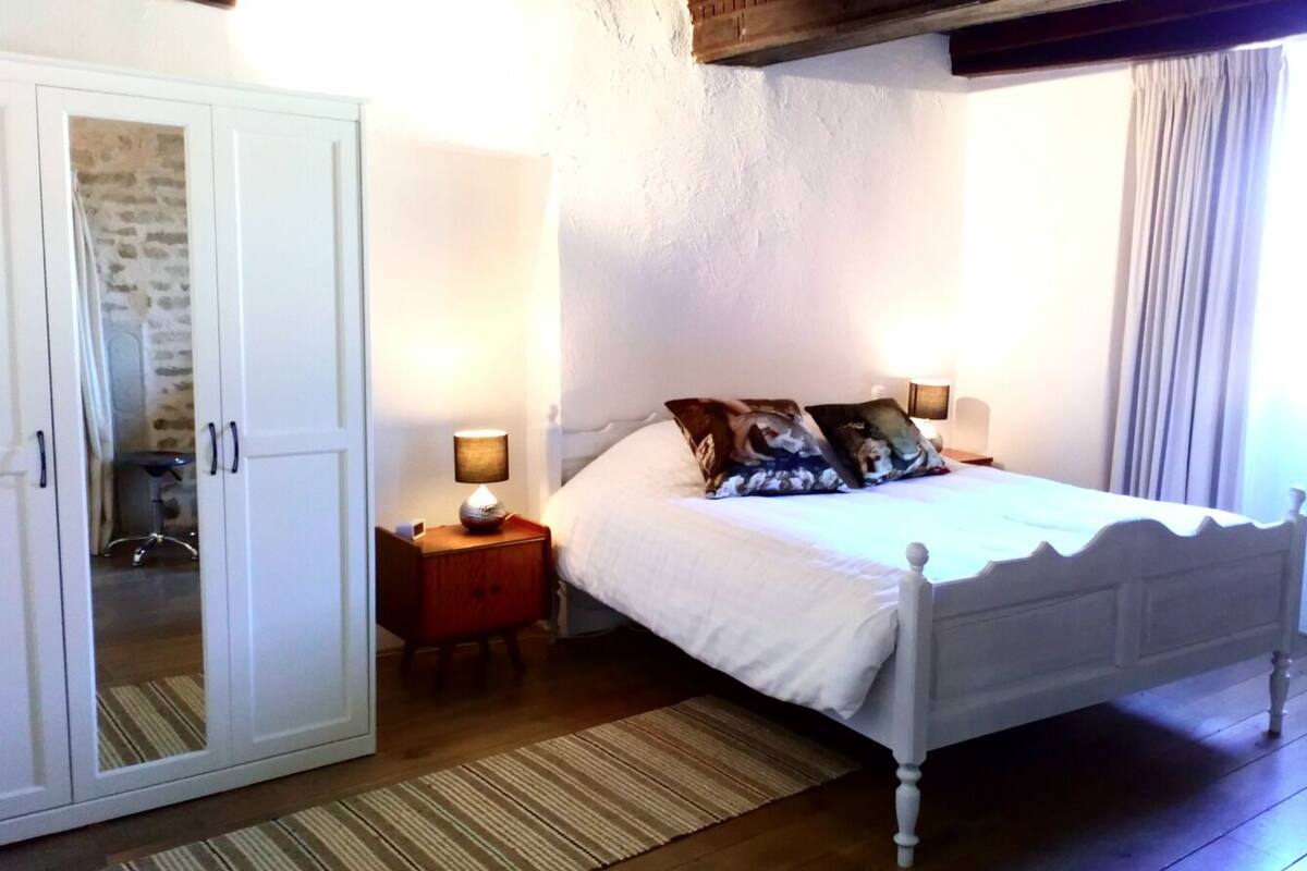 A bedroom with a white bed, a wooden wardrobe, a nightstand with a lamp, and a window with curtains.