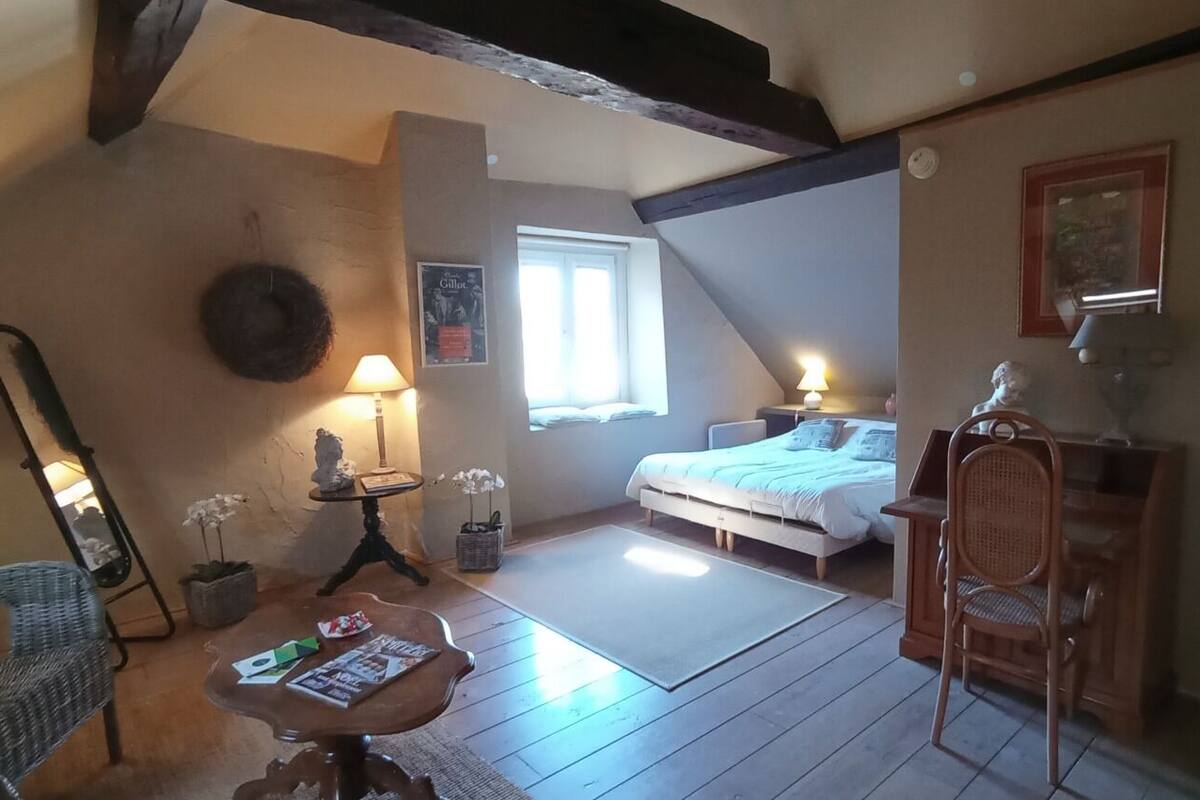 A cozy bedroom with a bed, a chair, a table with magazines, a lamp, and a window.