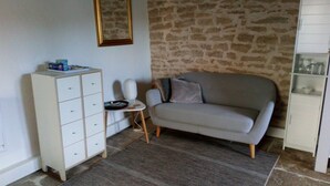 A room with a stone wall, a white cabinet, a grey sofa, and a small table with a lamp. - La Vallée Verte (Grandchamp)