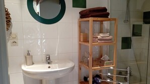 A bathroom with a white sink, a wooden shelf with towels, a mirror, and a glass shower enclosure. - La Vallée Verte (Grandchamp)