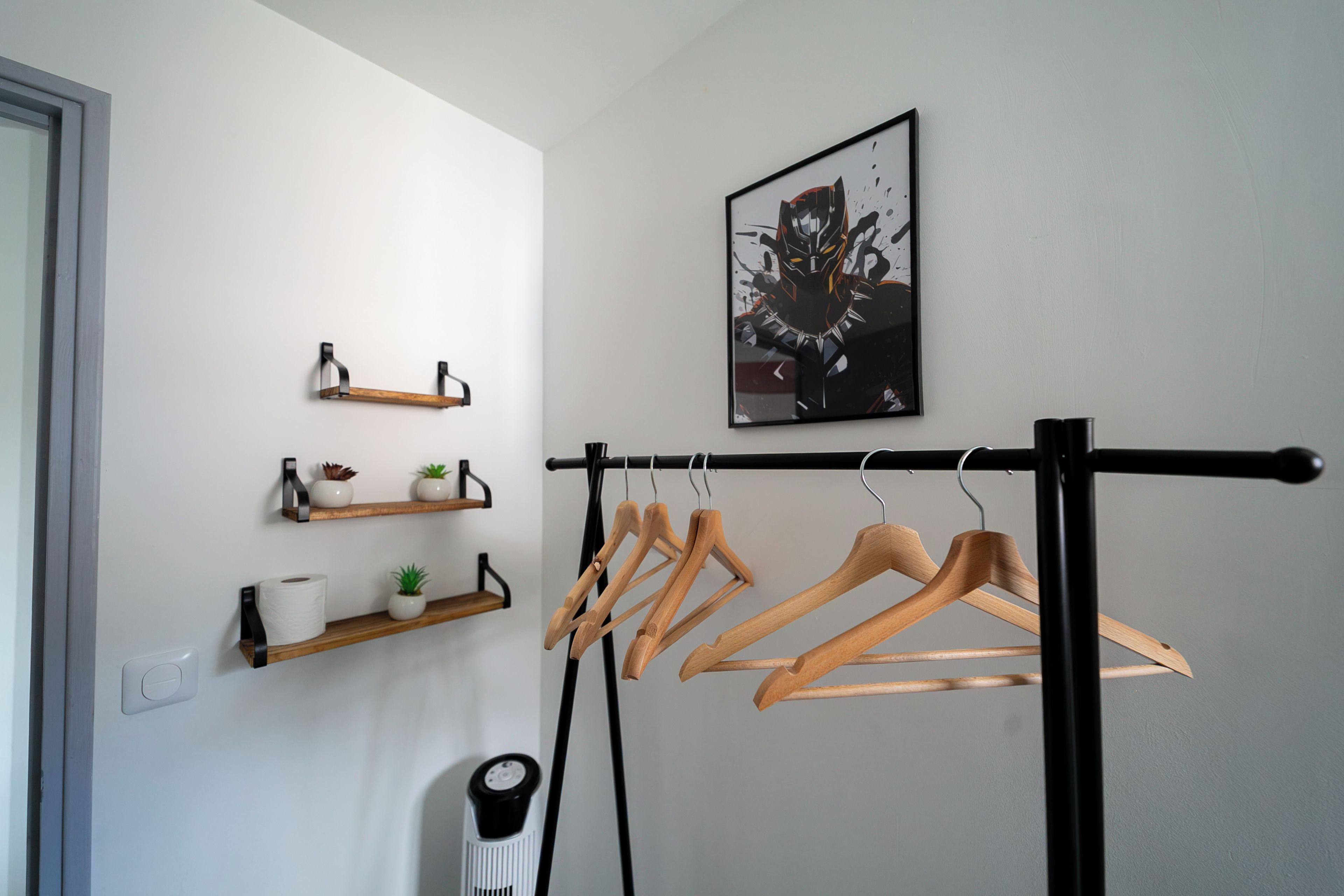 A room with a black metal drying rack, wooden shelves with decorative items, and a framed artwork on the wall.