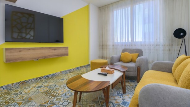 A living room with a yellow wall, a sofa, a coffee table, and a TV mounted on the wall. - Yelloscape/ Gemmayze (Beirut)