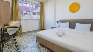 A hotel room with a bed, a desk with a chair, a window with curtains, and a patterned floor. - Yelloscape/ Gemmayze (Beirut)