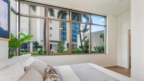 Un dormitorio moderno con un ventanal grande, una cama con ropa de cama estampada y vistas a un edificio y palmeras afuera.