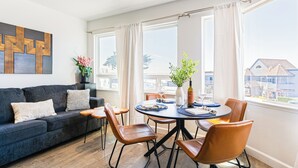 Ett modernt vardagsrum med en mörkblå soffa, ett runt matbord med stolar och stora fönster som ger utsikt över byggnaderna.