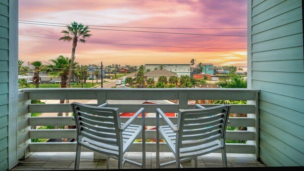A balcony with two white chairs and a table, overlooking a residential area with houses and palm trees under a sunset sky. - Sleeps 33! 4 Condos, Walk to the Beach, Resort-Style Pool, Endless Summer Resort (Port Aransas)