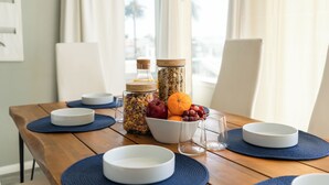 Een houten eettafel met witte borden, blauwe placemats en een schaal fruit en noten.