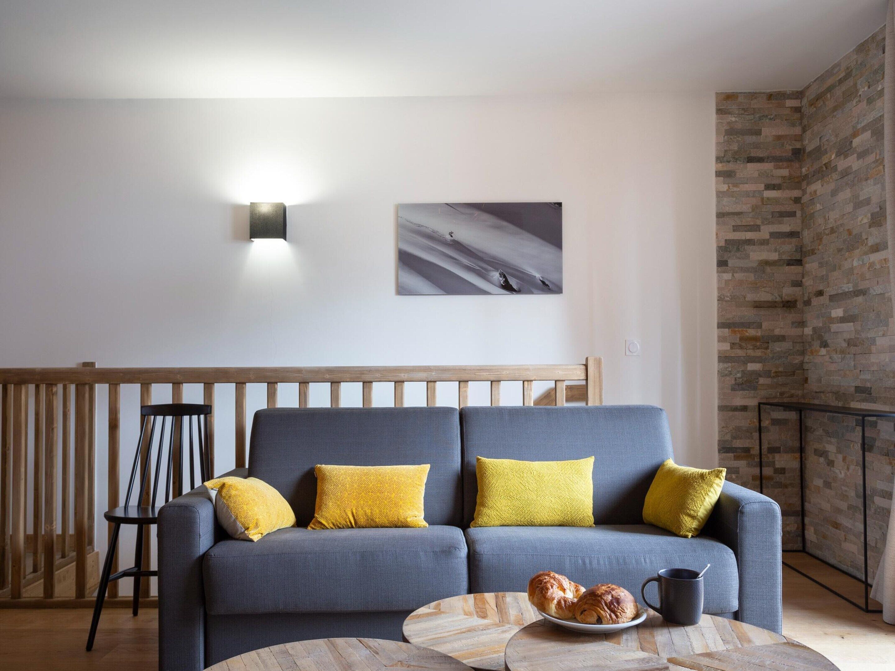 A modern living room with a grey sofa, yellow cushions, a wooden coffee table with croissants, and a wall-mounted light.