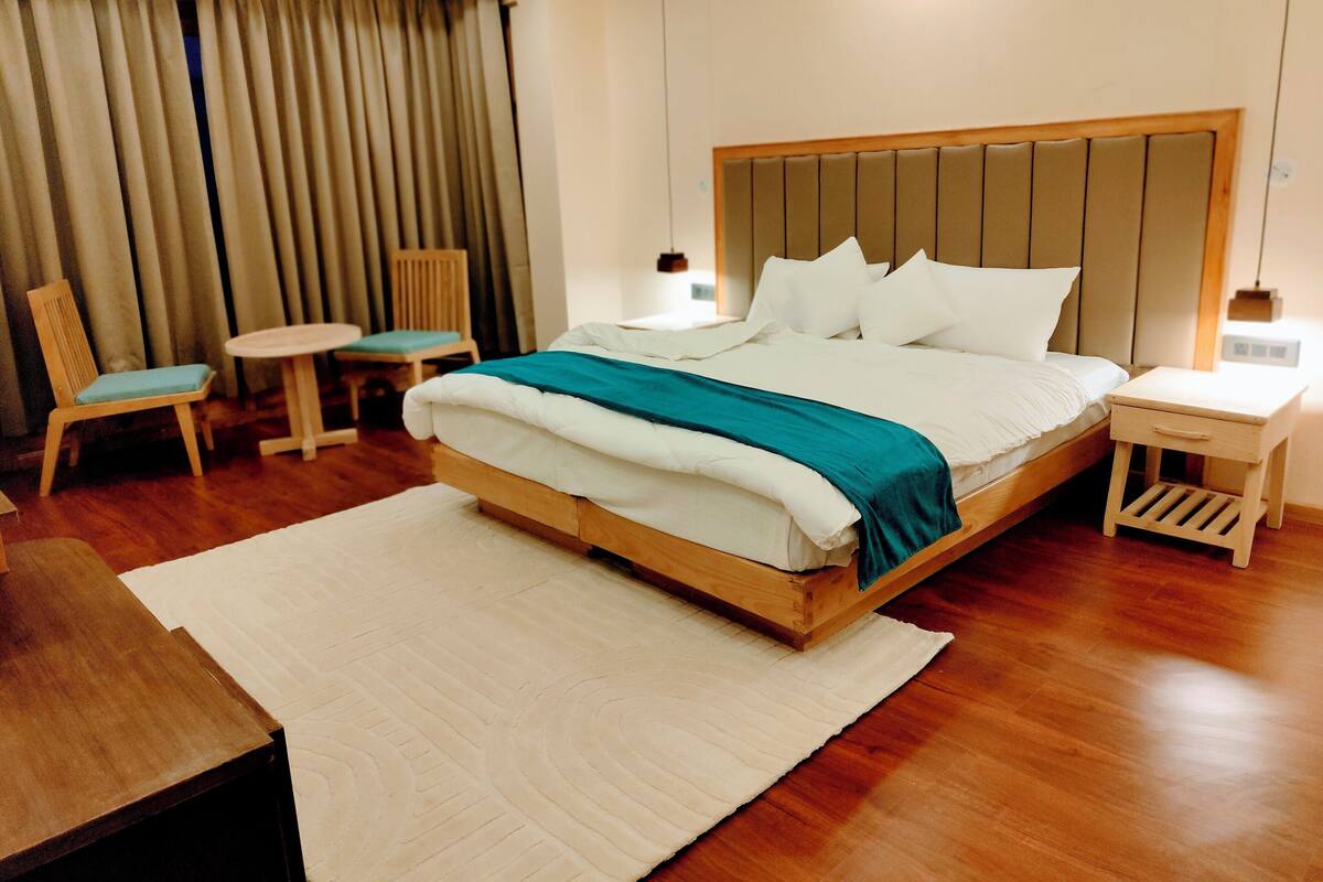A hotel room with a large bed, a wooden nightstand, a small table, a chair, and a large window with curtains.