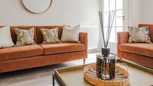 A living room with two brown sofas, a round mirror, and a wicker coffee table with a glass vase. - Skye Sands | Kinburn (No 2a) (St Andrews)
