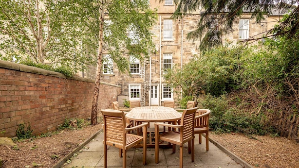 An outdoor patio with a round wooden table and chairs, surrounded by a brick wall and dense foliage. - Skye Sands | Kinburn (No 2a) (St Andrews)